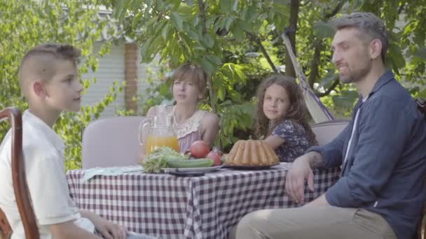 Tata, Mama i ich dwoje dzieci siedzą przy stole, jedzą lunch w ogrodzie, ciesząc się słoneczny dzień. Rodzina patrząc w aparat uśmiechnięty. Summertime, szczęśliwa Rodzina — Wideo stockowe