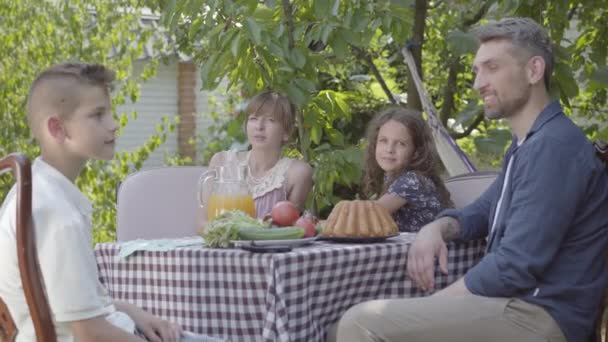 Papa, maman et leurs deux enfants assis à une table, déjeunant dans le jardin en profitant d'une journée ensoleillée. Famille regardant dans la caméra souriant. L'été, famille heureuse — Video