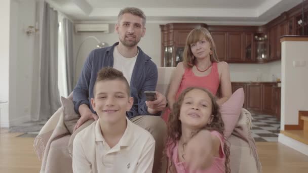 Joven familia alegre viendo la televisión mientras están sentados juntos en casa. El padre sostiene el control remoto y cambia los canales, la esposa se sienta al lado de su marido y dos hijos están sentados con sus padres — Vídeo de stock