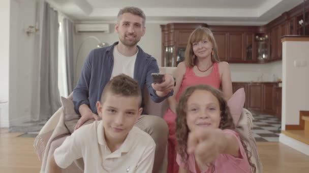 Portret młodej rodziny oglądania telewizji, siedząc w domu razem. Ojciec trzyma pilota i przełącza kanały, żona siedzi obok męża i dwoje dzieci siedzi z ich — Wideo stockowe