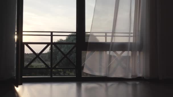 Μεγάλο ξύλινο ανοιχτό παράθυρο με κουρτίνες που χαιρετάνε τον άνεμο. Όμορφο καλοκαιρινό τοπίο πίσω από το μπαλκόνι. Η κάμερα κινείται δεξιά. Η ιδέα του ηλιοβασιλέματος — Αρχείο Βίντεο