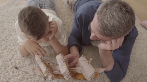 Portret schattige vader en zijn zoon liggen thuis op de vloer op het pluizige tapijt spelen met schip model close-up. Gezins tijd samen doorbrengen. Achteraanzicht — Stockvideo