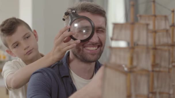 Retrato bonito criança segurando loupe em mãos olhando através dele nos detalhes do brinquedo. Pai e seu filho brincando com o modelo de navio close-up. O pai a ensinar o filho. Família passar tempo juntos — Vídeo de Stock