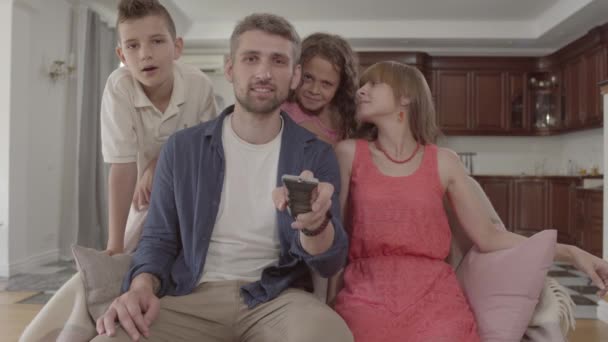 Retrato joven familia viendo la televisión en casa juntos. El padre tiene el control remoto y cambia los canales, la esposa se sienta al lado de su marido y dos hijos están con sus padres. Tiempo libre familiar . — Vídeos de Stock