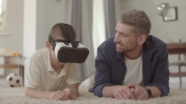 Porträt schöner Vater und Sohn, die mit Virtual-Reality-Brille auf dem Boden auf dem flauschigen Teppich liegen, glückliche, freundliche Familie zu Hause. Beziehung Vater-Sohn. — Stockvideo