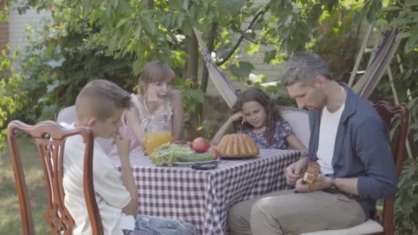 O pai toca guitarra para a família numa mesa de jantar no quintal. Relações familiares amigáveis . — Vídeo de Stock