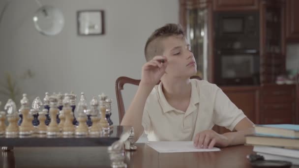 家のテーブルに座っている小さな男の子の肖像画。男は何かを考えてペンを持っている. — ストック動画
