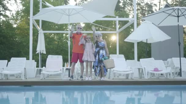 Portret szczęśliwy dojrzały para z mały wnuczka stojący przy the basen i fala ich ręce przy the aparat fotograficzny. Babcia i dziadek spoczywają na wnuczku. Rodzina przyjazna dla przyjemności. — Wideo stockowe