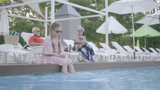 背景のプールの近くのサンベッドに横たわっている老夫婦。水の中で彼女の足でプールの端に座っているピグテールを持つ小さな面白い女の子。幸せなフレンドリーな家族。ホテルで休む — ストック動画