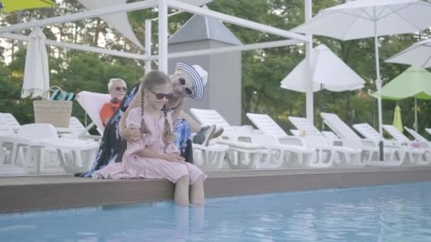 Großmutter umarmt ihre Enkelin am Pool sitzend und schaut in die Kamera. Großvater ruht auf einer Liege im Hintergrund. — Stockvideo