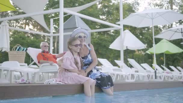 Schattige kleine grappig meisje met Pigtails en volwassen vrouw zittend op de rand van het zwembad met hun voeten in het water terwijl senior man liggend op zonnebank op de achtergrond. Gelukkig vriendelijke familie. Rusten in — Stockvideo