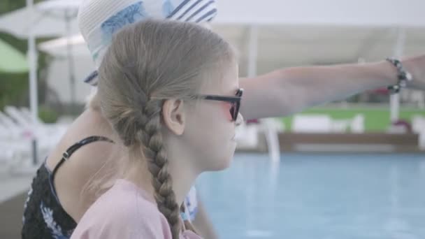 Klein schattig meisje met staartjes en volwassen vrouw dragen zonnebril zittend op de rand van het zwembad en praten. Rust en vrije tijd van grootmoeder en kleindochter. Gelukkige vriendelijke familie. — Stockvideo