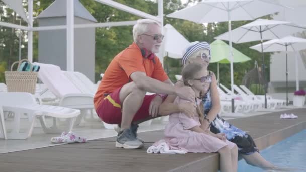 Happy mature couple on the edge of the pool hugging little granddaughter. Grandmother, grandfather and grandchild waving hands. Happy friendly family. Rest in hotel. Recreation and leisure outdoors — Stock Video