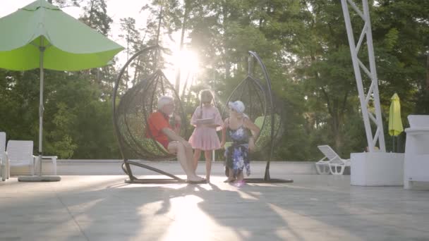 Kis aranyos lány Pigtailek visel nyári ruha és napszemüveg áll a tabletta mellé nagyszülei ült lógó székek. — Stock videók