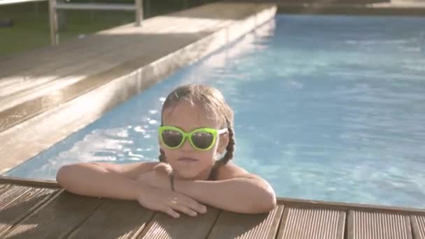 Retrato chica bastante divertida en gafas de sol amarillas mirando en la cámara sonriendo, mirando fuera de la piscina, aferrándose al borde. Niño divirtiéndose en el complejo hotelero. Recreación y ocio al aire libre . — Vídeo de stock