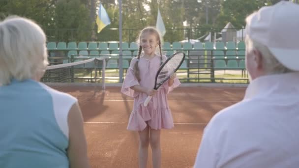 Schattig grappig meisje met twee Pigtails spelen tennis buiten terwijl haar grootouders zitten in de buurt van haar te steunen. Heerlijk kind het verhogen van een racket. Recreatie en vrije tijd buitenshuis. — Stockvideo