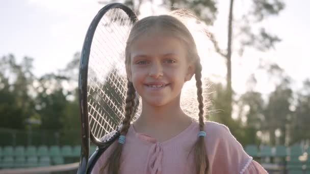 피그테일과 테니스 라켓을 어깨에 대고 여름 햇살을 쬐고 서 있는 카메라를 바라보며 귀여운 미소 짓는 소녀를 초상화합니다. 레크리에이션 및 레저 아웃도어. — 비디오
