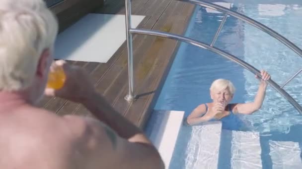 Vista posteriore dell'uomo maturo che beve succo guardando sua moglie, nuotando in piscina. Coppia matura che si rilassa in piscina nel complesso alberghiero insieme. Felice famiglia amorevole. Riposo in hotel — Video Stock