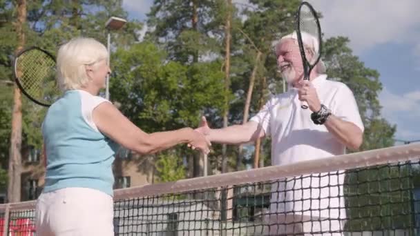 Улыбающаяся зрелая пара пожимает руку после игры в теннис на теннисном корте. Активный отдых на свежем воздухе. Старший мужчина и женщина веселятся вместе — стоковое видео
