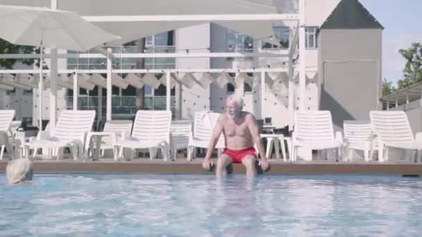 Boldog érett pár relaxáló a szálloda medencéjében együtt. Aranyos idős férfi ül a medence szélén, míg nő úszunk a vízben. Boldog barátságos család. Pihenés a hotelben — Stock videók