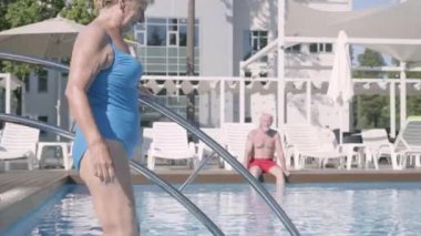 Kocası arka planda otururken havuza yürürken çekici yaşlı kadın. Otel kompleksindeki havuzda dinlenen olgun çift. Mutlu sevgi dolu bir aile. Otelde dinlenin