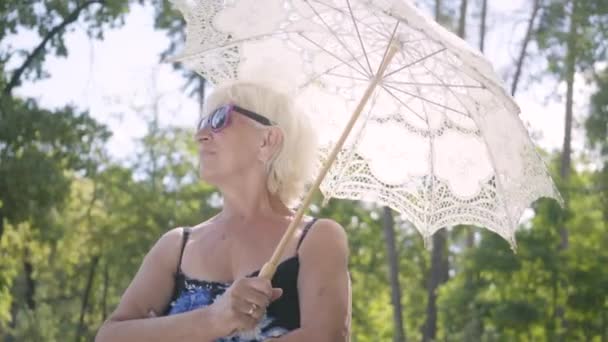 Портрет позитивно улыбающейся зрелой женщины в солнечных очках, стоящей на открытом воздухе под белым зонтиком. Старший мужчина встречается с женой в парке, целуется. Отдых на свежем воздухе в жаркий солнечный день — стоковое видео