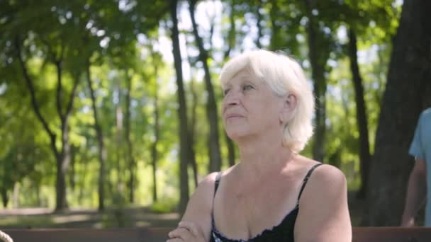 Retrato de una atractiva mujer madura sentada en el banco del parque. Hombre mayor sonriente viniendo detrás de ella y cubriendo sus ojos con sus manos. Viejo y mujer tienen una cita — Vídeos de Stock