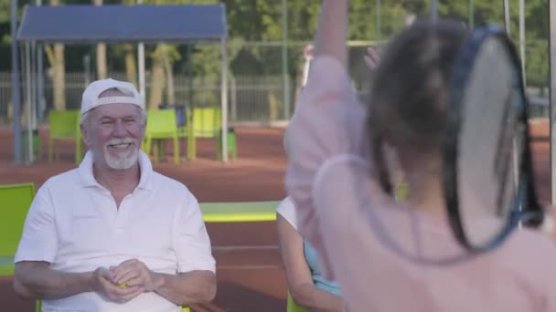 Achteraanzicht van schattig grappig meisje met twee Pigtails staande op de tennisbaan Holding racket, zwaaien hand aan grootmoeder en grootvader. Gelukkige familie die lucht kusjes naar elkaar stuurt. Actieve vrijetijdsbesteding — Stockvideo