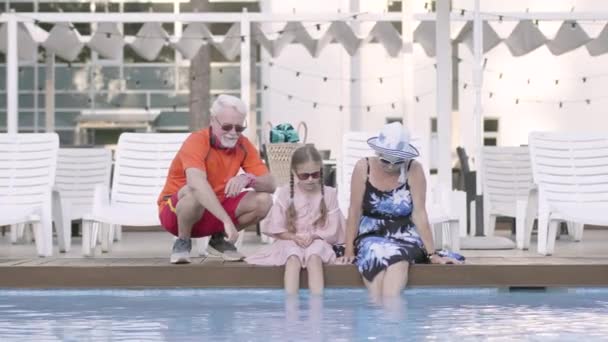 Feliz pareja madura y su nieta sentada en el borde de la piscina. Abuela, abuelo y nieto descansando juntos. Feliz familia amistosa. Vista frontal — Vídeos de Stock