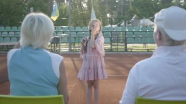 Tenis kortu raket tutan duran iki pigtails ile sevimli komik kız, kameraya sırtlarını ile oturan büyükanne ve dedesi, hava öpücük gönderme. Yaz tatili