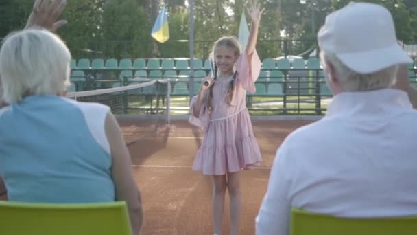 Drăguţă fetiţă zâmbitoare şi fericită, cu o rachetă de tenis în mâini stând pe terenul de tenis uitându-se în cameră. Bunicul și bunica se bucură de mâinile nepoatei — Videoclip de stoc