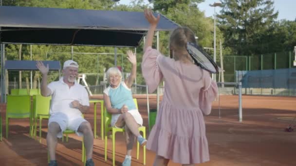 Gadis kecil yang lucu tersenyum bahagia dengan raket tenis di tangannya berdiri di lapangan tenis melihat ke kamera. Kakek, nenek dan gadis bersukacita di tangan gelombang cucu mereka — Stok Video