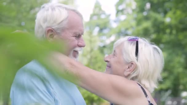 Позитивная зрелая пара целуется и смотрит в камеру улыбаясь. Старшая женщина обнимает своего мужа. Прекрасная счастливая семья отдыхает на свежем воздухе. Летний отдых. Романтическое свидание — стоковое видео