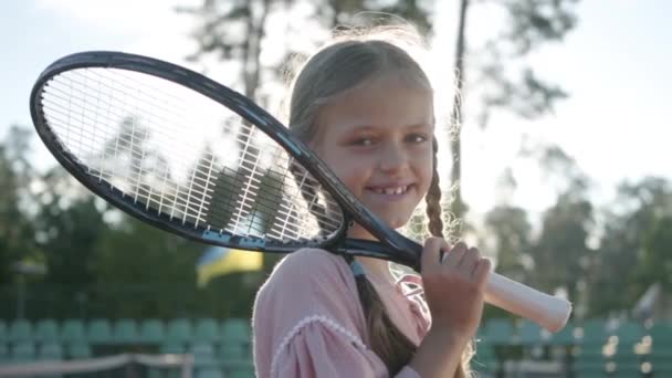 ピグテールと彼女の肩にテニスラケットを持つかわいい小さな笑顔の女の子は、夏の太陽の光の中に立ってカメラを見て、屋外でのレクリエーションやレジャー。スローモーション. — ストック動画