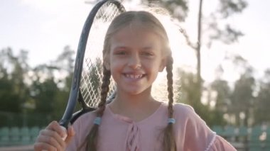 Portre sevimli küçük gülümseyen kız pigtails ve omzunda bir tenis raket ile kamera içine yaz güneşi ışınları ayakta bakarak. Açık havada eğlence ve eğlence.