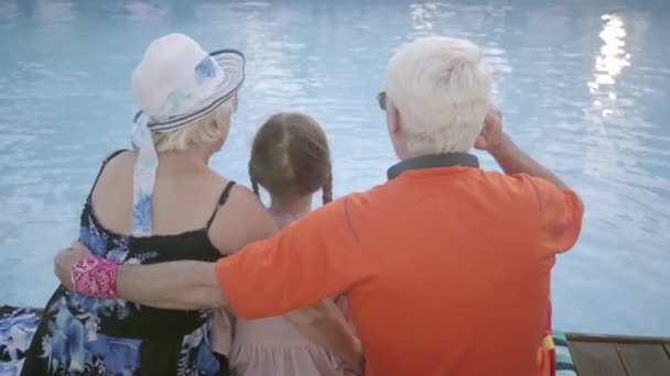 Зрелый мужчина, женщина и маленькая девочка сидят на краю бассейна, вид сзади. Бабушка, дедушка и внук отдыхают у воды. Счастливой дружеской семьи. Отдых в гостинице — стоковое видео