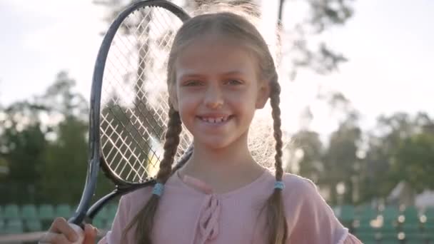 Retrato linda niña sonriente con coletas y una raqueta de tenis en el hombro mirando a la cámara de pie en los rayos del sol de verano. Recreación y ocio al aire libre . — Vídeo de stock