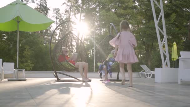 Old couple sitting on a hanging chair on the sunset, relaxing in the hotel complex together. Cute little girl running to grandparents, the man giving her a tablet. Recreation and leisure outdoors. — Stock Video