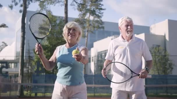 Linda pareja madura preparándose para jugar al tenis en la cancha de tenis. La mujer sosteniendo una raqueta y una pelota a punto de pasar. Ocio activo al aire libre. Hombre y mujer mayores divirtiéndose juntos — Vídeo de stock