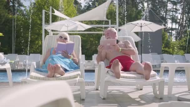 Жизнерадостная позитивная пара, лежащая на санках возле бассейна. Женщина читает книгу, в то время как мужчина работает с планшетом. Счастливая любящая семья отдыхает вместе. Отдых в отеле. Вид спереди — стоковое видео