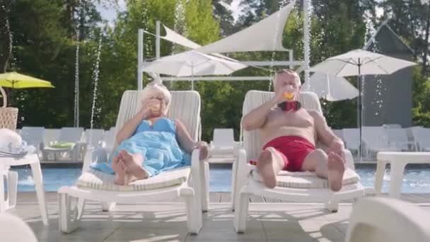 Pareja madura tumbada en tumbonas cerca de la piscina bebiendo jugo, hablando y sonriendo. Feliz familia amorosa. Recreación y ocio al aire libre . — Vídeo de stock