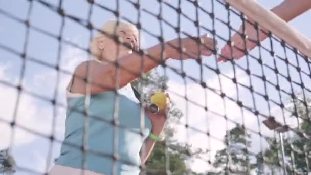 Η ενήλικη γυναίκα κάνει χειραψία με έναν άγνωστο αντίπαλο που στέκεται σε ένα γήπεδο τένις στις ακτίνες του καλοκαιριού ήλιου. Αναψυχή και αναψυχή σε εξωτερικούς χώρους. — Αρχείο Βίντεο
