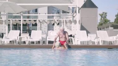 Mutlu olgun çift birlikte otel kompleksinde havuzda rahatlatıcı. Kadın suda yüzerken havuz kenarında oturan sevimli yaşlı adam. Mutlu dost bir aile. Otelde dinlenin