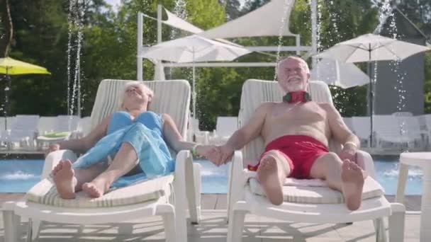 Anziani coppia positiva sdraiata su lettini vicino alla piscina tenendosi per mano e sorridendo. Felice famiglia amorevole. Riposa in albergo. Vista frontale — Video Stock