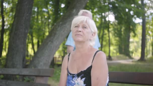 Portrait of adorable mature woman sitting on the bench in the park. Smiling senior man coming up behind her and covering her eyes with his hands. Old man and woman have a date — Stock Video