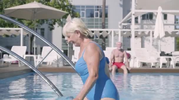 Glückliches älteres Paar, das sich gemeinsam am Pool der Hotelanlage entspannen kann. attraktive Seniorin sonnt sich, kommt aus dem Pool und winkt ihrem Mann zu. glückliche freundliche Familie. Erholung im Hotel — Stockvideo