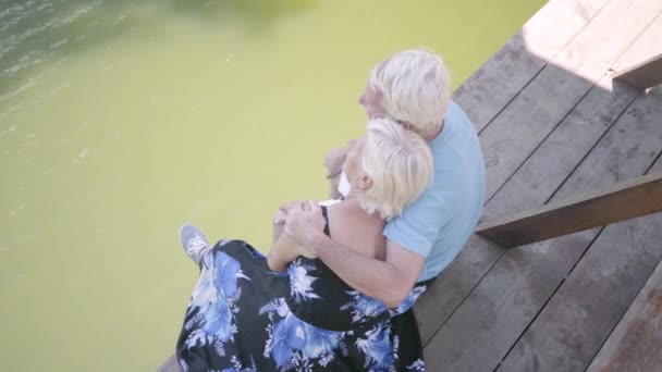 Vista superior de um casal maduro feliz sentado no banco perto do rio, admirando a natureza. A senhora a beijar o marido. Lazer ao ar livre. Câmera em movimento direito — Vídeo de Stock