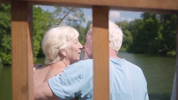 Šťastný dospělý pár sedí na lavičce u řeky, obdivuje přírodu. Starší žena líbala svého manžela. Volný čas venku. — Stock video