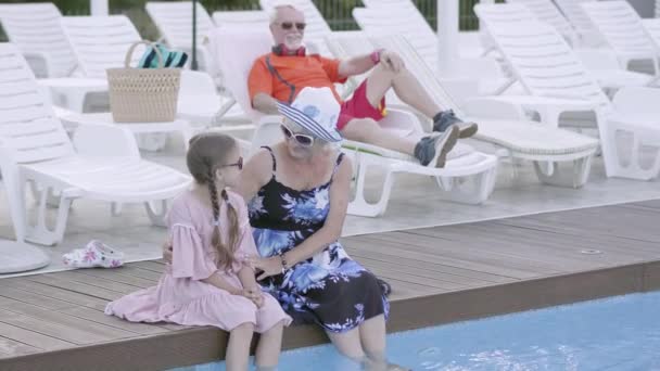 La abuela abraza a su nieta sentada junto a la piscina y mira a la cámara. Abuelo está descansando tumbado en una tumbona en el fondo . — Vídeo de stock