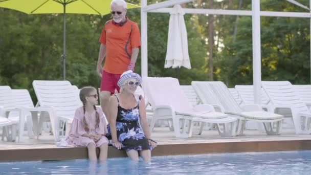 Trochę cute girl z warkoczami i Starsza kobieta siedzi na skraju basenu z nogami w wodzie. Dziadek joing nich. Miłej rodziny. Spoczywaj w hotelu — Wideo stockowe